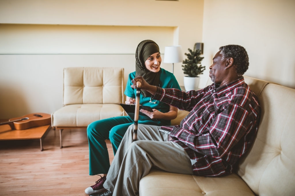 A Muslim female doctor is visiting her older patient of Afro-American ethnicity. An Arabian female healthcare worker is helping her older male patient in the living room at home.